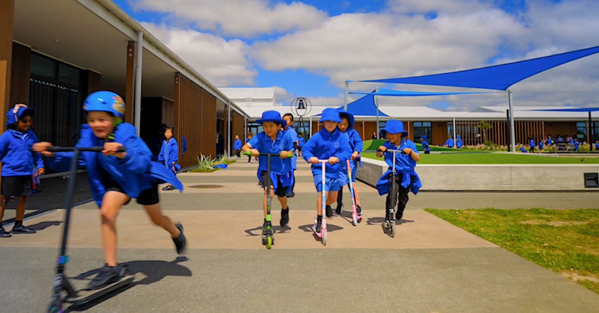 wigram primary students in blue and blue bucket hats riding scooters on paving at school