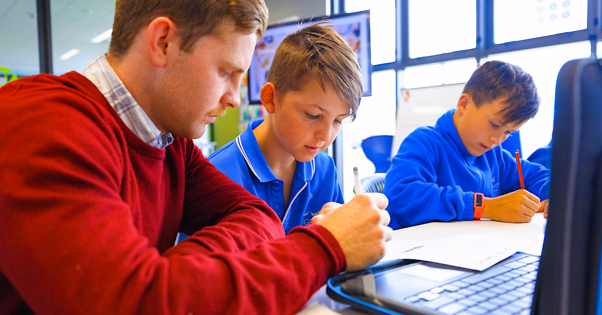 two pupils in blue with teacher in red jumper working at a desk with laptop and pens