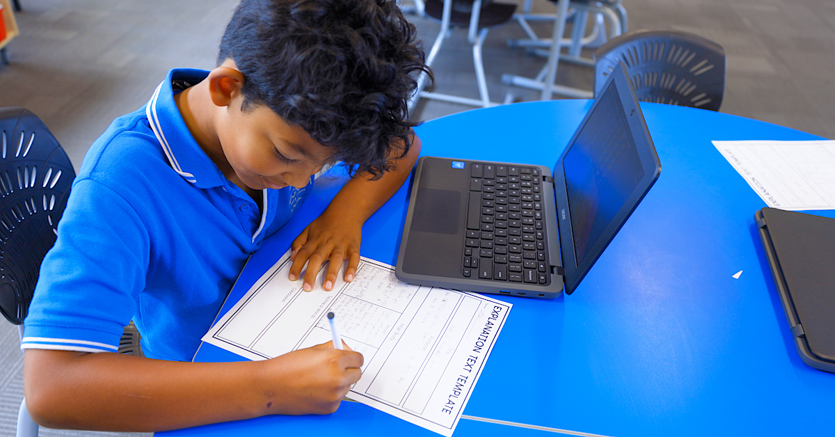 young male pupil at a blue desk with laptop writing on a white page